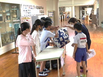校舎の広い廊下に設置された長テーブルに6名の生徒たちが横一列に立ち、体育服を着た男の子と母親の親子に大きな袋を手渡している写真