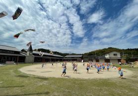 園庭の空になびいている鯉のぼりと園庭を走り回る園児たちの写真
