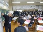 教室で、教壇に立ち黒板を指さす職員と、それを立ってみている児童の写真