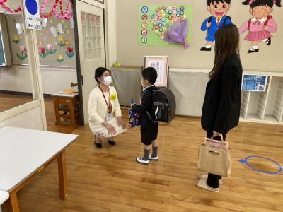 教室で新一年生の目線に合わせてしゃがんで話をする先生と子供とそれを見ている保護者の写真