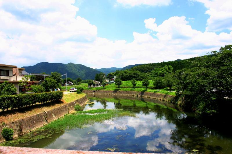 奥に山並みが見える手前に、民家や林に挟まれた川が流れている写真