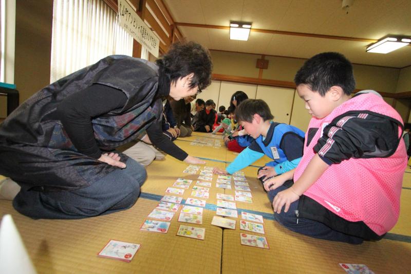 右側にピンク色と青色のゼッケンをつけた2名の男の子、左側に大人の女性が向かい合って座りかるた大会を行っている様子の写真