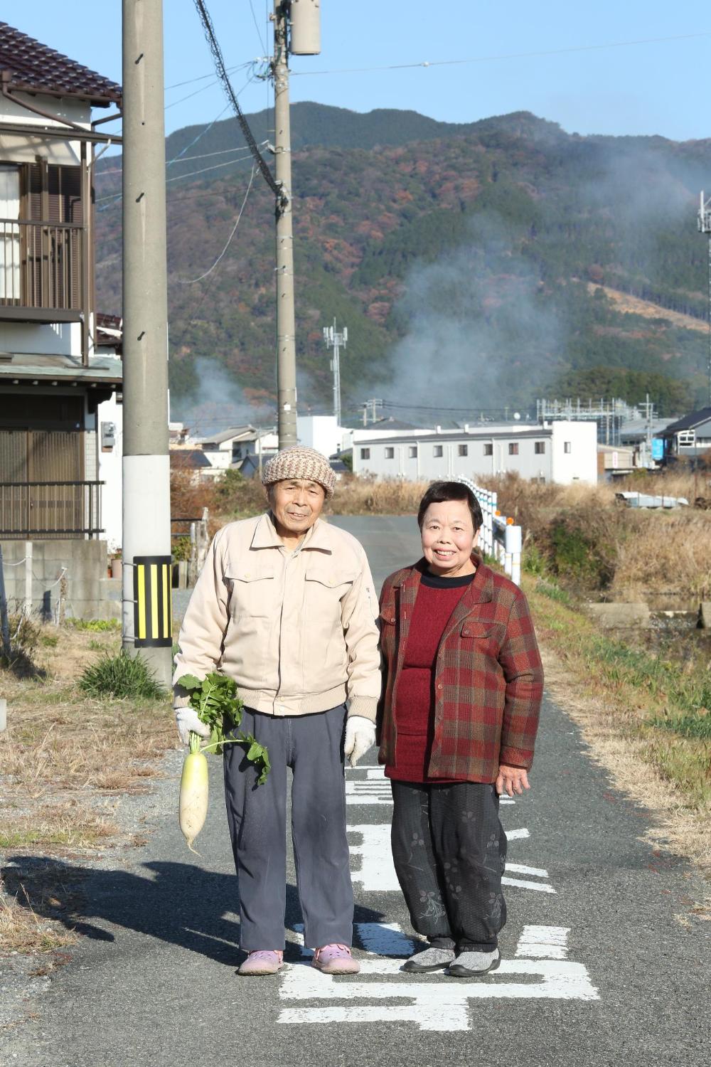 城戸 武さんと城戸 政子さんが大根を持ち、あぜ道で寄り添う写真