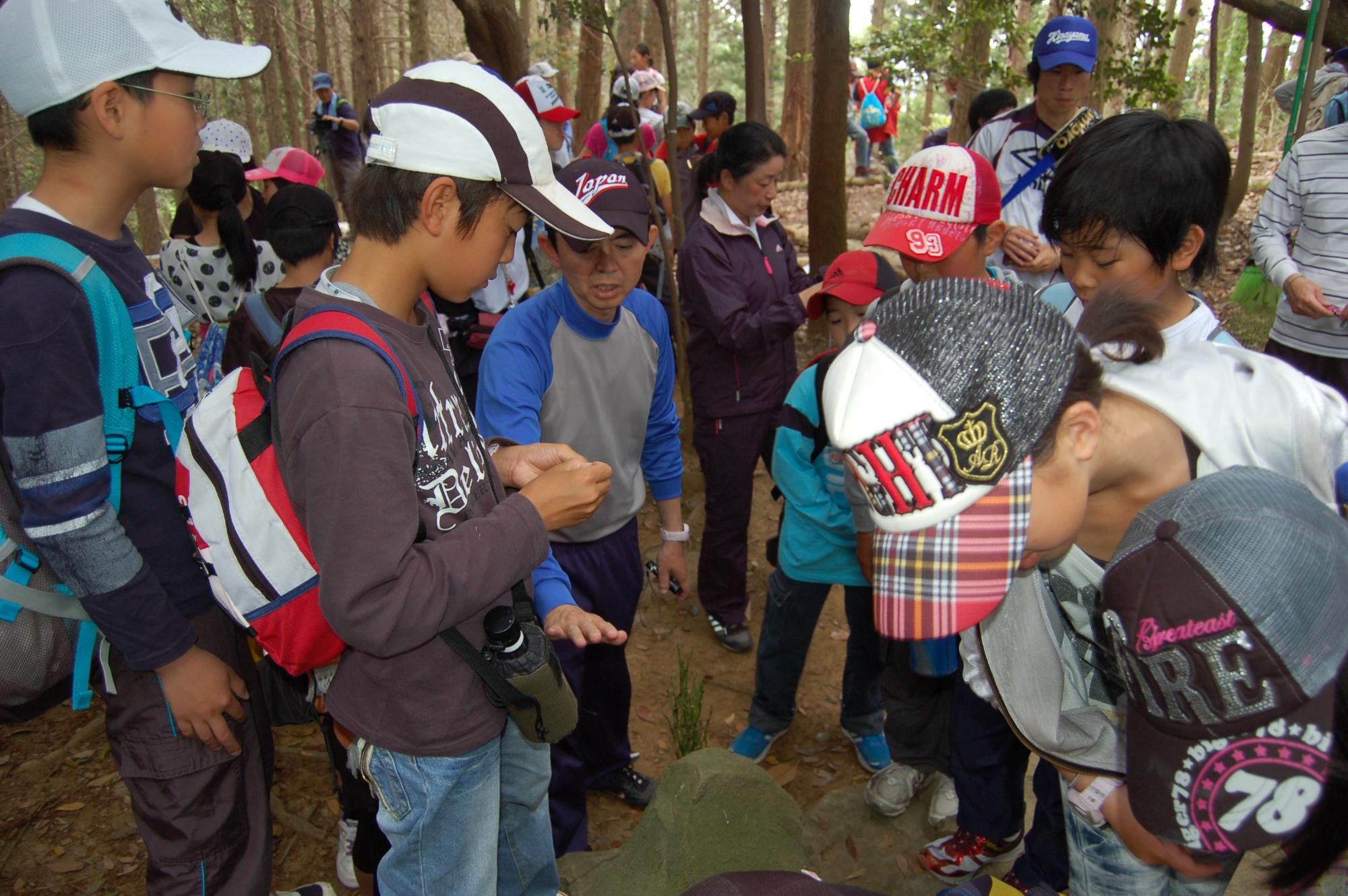 木が生い茂る山の中にリュックを背負って帽子をかぶった子供たちがたくさん写っている写真
