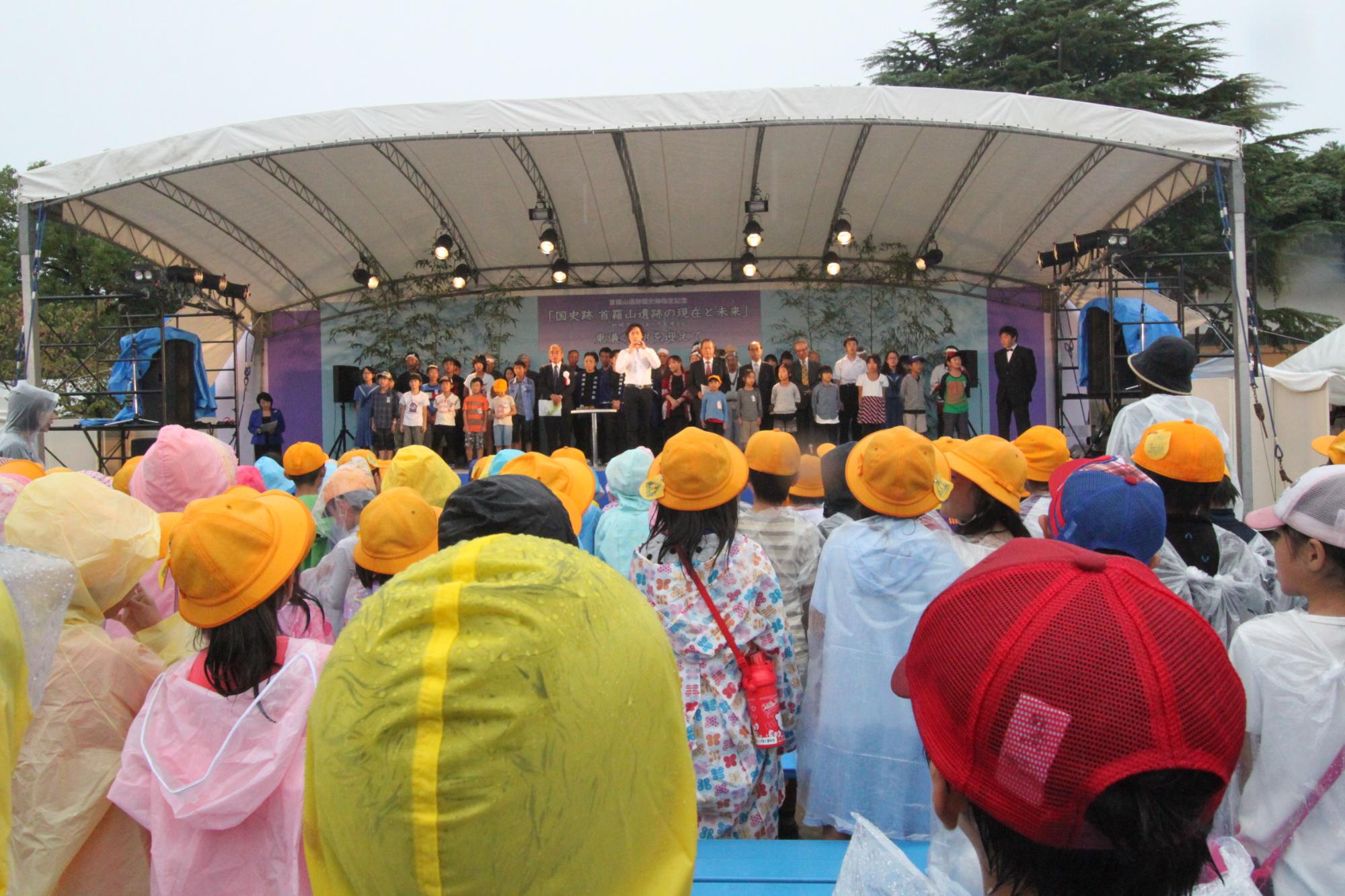 「国史跡 首羅山遺跡の現在と未来」と書かれた屋外ステージ中央で東儀秀樹さんが演奏しているのをステージ上の大人や子供たちや、ステージの下で帽子をかぶってカッパを着た子供たちがそれを見ている様子の写真