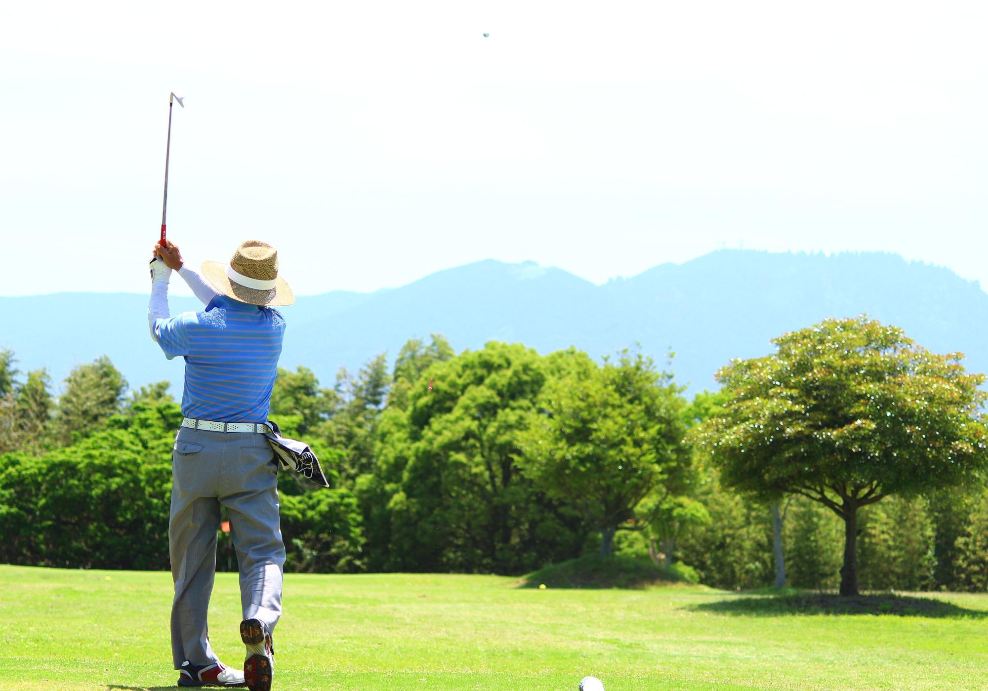 自然の中で帽子をかぶり青いウェアを着た男性が、ゴルフクラブをスイングしている写真