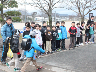 挨拶運動のため並んで立っている地域の方々や小学生の子供たちと登校中の小学生2人の写真