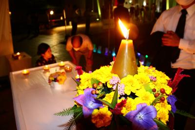 白いテーブルの上に置かれた台に黄色や紫の花が飾られており、その中央にロウソクが飾られている写真