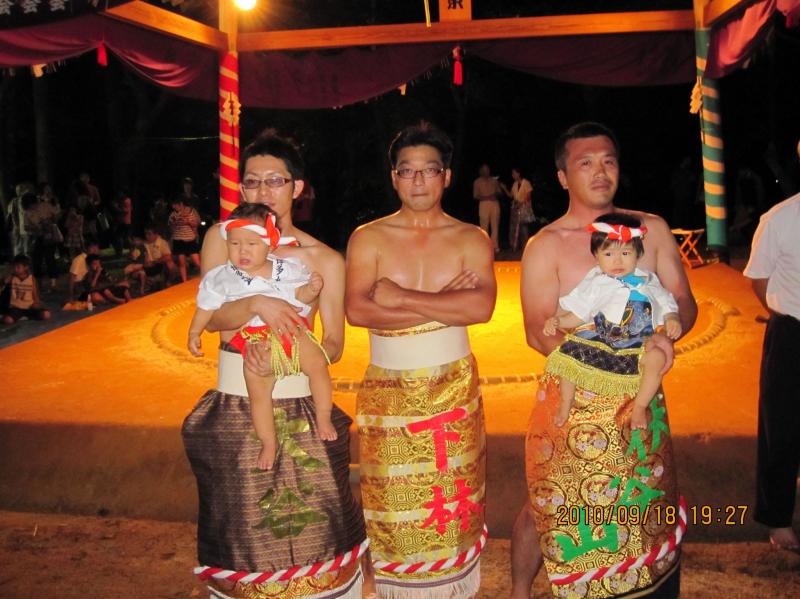 化粧まわしをしめた3人の男性が土俵の前に並んで、中央の男性は腕を組み両隣の男性は子供を抱っこしている写真