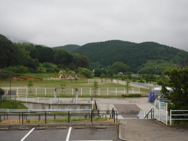 手前の駐車場の階段を下りた先に芝生の広場があり、その奥には山が見える写真