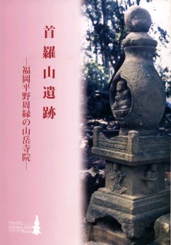 右半分に薩摩塔、左半分にピンク色の背景に赤字で「首羅山遺跡―福岡平野周縁の山岳寺院―」と書かれた表紙の画像