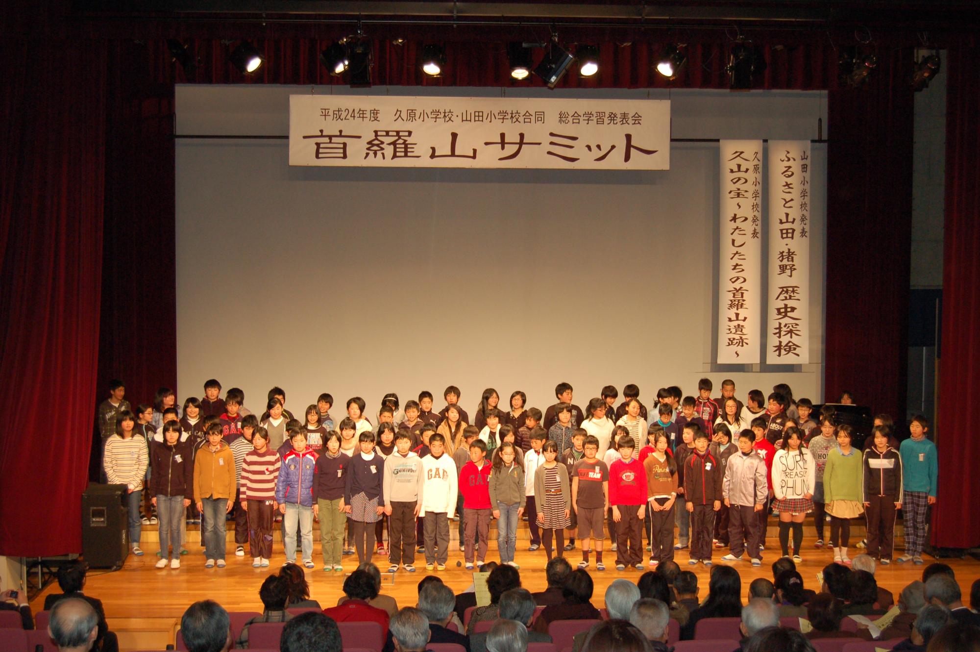「首羅山サミット」と書かれた屋内のステージで並んで発表する子供たちと、客席で見ている大人達の写真