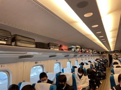 新幹線の荷物棚にスーツケースが入れられ、学生達が2列の座席に座っている新幹線車内の写真