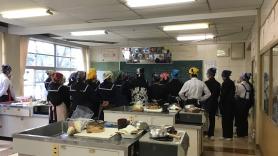 調理室の前方に集まっているエプロンや三角巾を付けた中学生たちの後ろ姿の写真