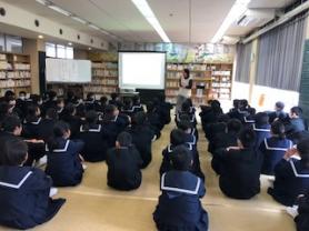 広い図書室のような場所で生徒たちが並んで床に座り、前方に設置されたプロジェクタースクリーンの横で話をしている才田衣恵先生の話を聞いている写真