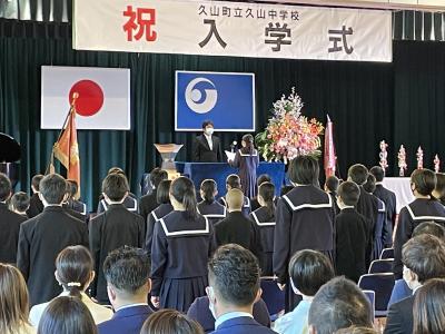 祝入学式と書かれた幕が掲げられた舞台で誓いの言葉を述べている女子生徒、演壇にたっている校長先生、起立している生徒たちの写真
