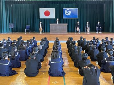 日の丸国旗と校章が掲げられた舞台の演壇で話をしている先生、椅子に座っている4名の先生、床に座って話を聞いている生徒たちの写真