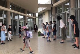 6年生の生徒たちと女性の教師が昇降口に横一列に立ち、登校してくる子供たちと挨拶を交わしている写真