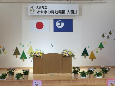 天井から「久山町立けやきの森幼稚園 入園式」と書かれた舞台看板、壇上に演題や鉢植えの花が並べられている写真