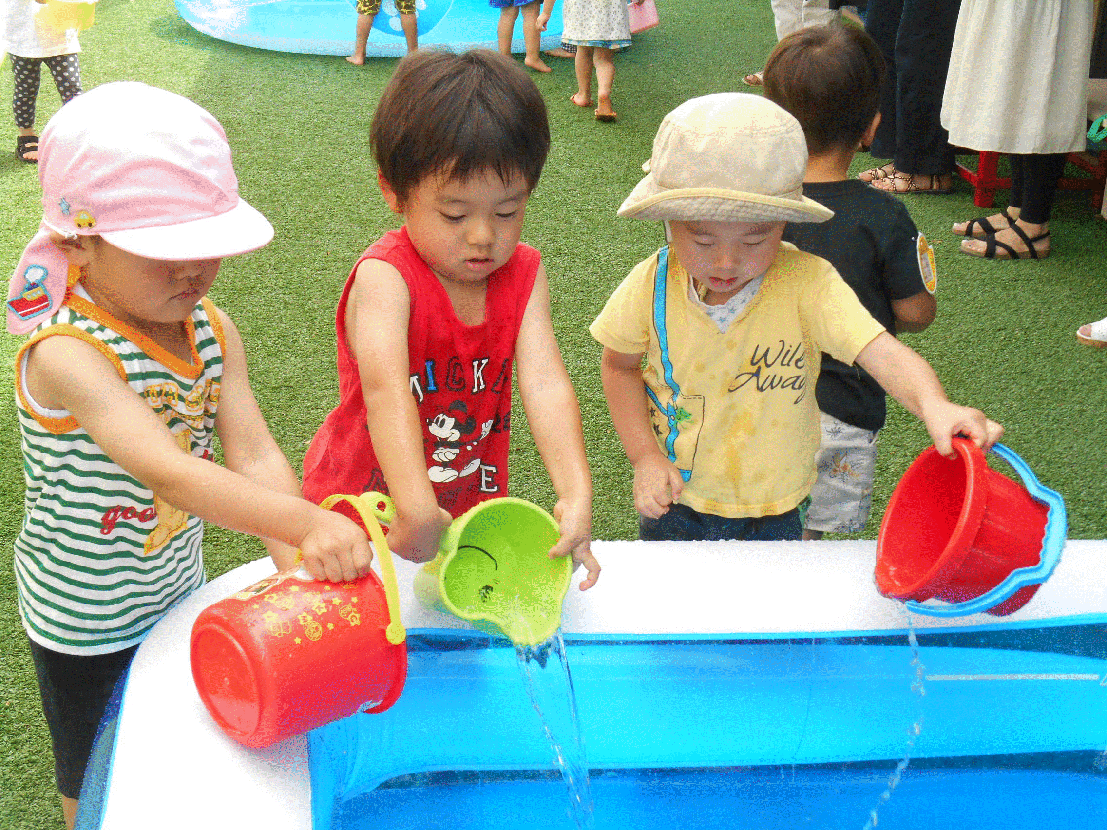 小さな子供たちが手に持ったバケツの水を大きなビニールプールに注いでいる写真