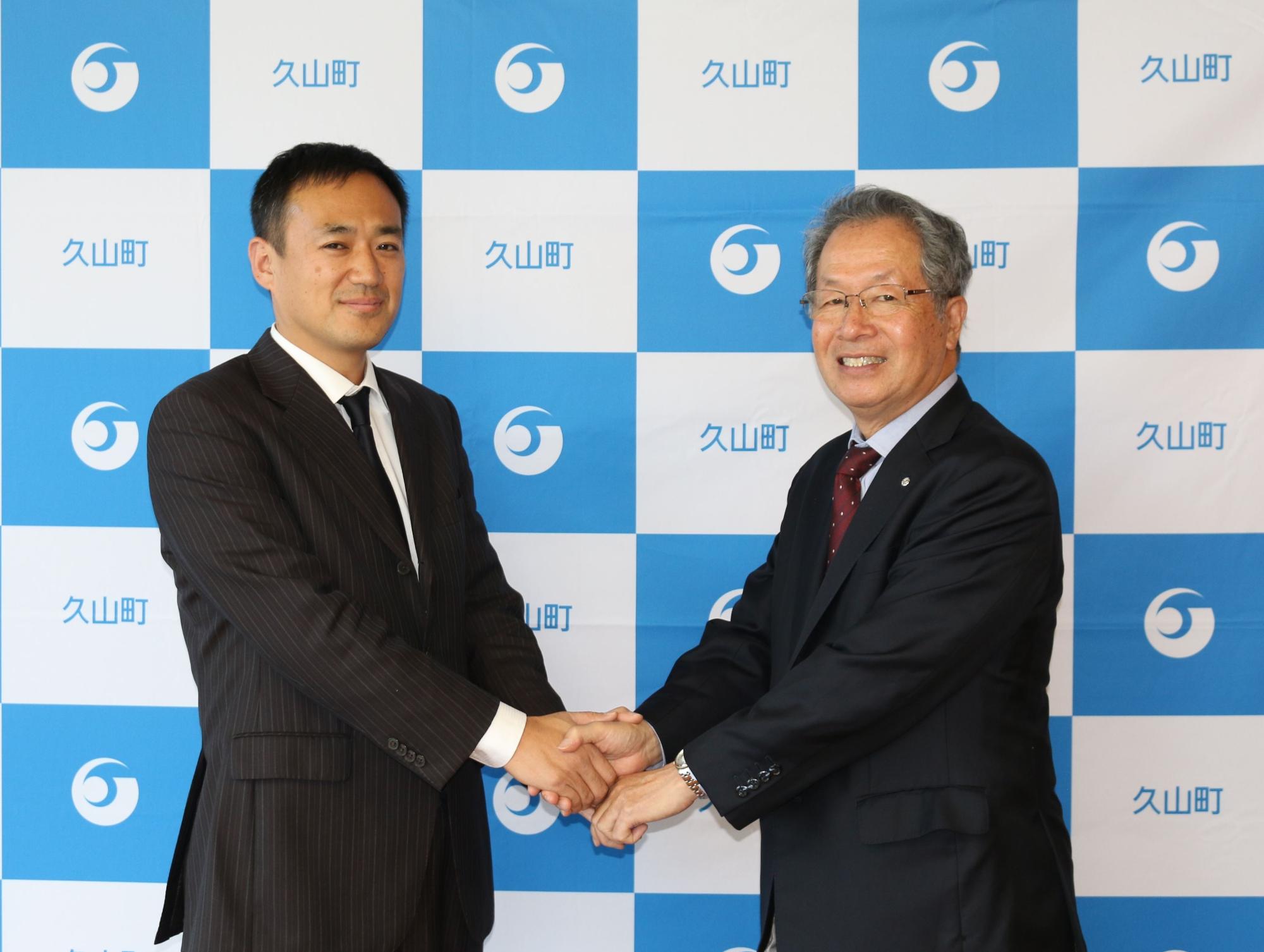 九州大学馬奈木教授と久芳菊司町長が両手で握手をしている写真。