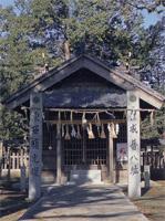 木々に囲まれた社殿の手前に、文字が刻まれた2本の石柱が立っている写真