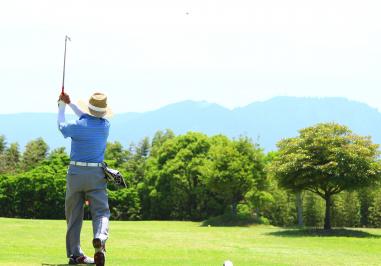 霞みがかった奥に山並みが見える自然豊かなゴルフ場で、麦わら帽子を被った男性がスイングを行っている写真