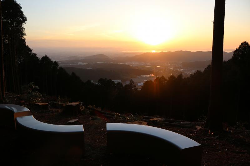 波型のベンチが設置された山頂から見下ろした街並みの先に、太陽が半分遠くの山に隠れている写真