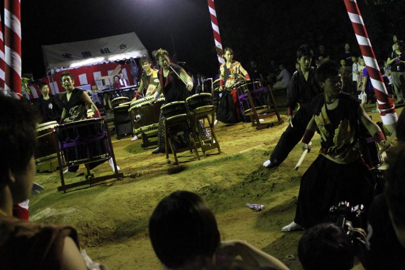 周囲で観客が見ているなか、日が暮れてライトアップされた屋外の祭りで和太鼓を演奏している人達の写真