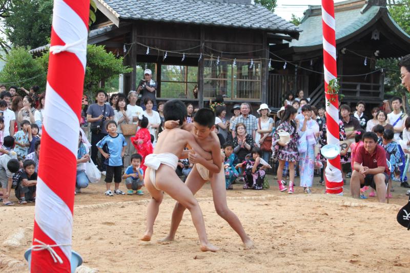 神社の境内で周囲に観客が見守るなか、中央の土俵で相撲をとっている2名の男の子の写真