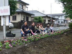 道路横の少し咲いてきている花々の後ろで7名の景観づくり団体の皆さんが座って記念撮影をしている写真
